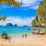 Wooden boats and tourists on a sandy beach in Thailand with small islands in the distance, part of the Thailand Classic Getaway tour